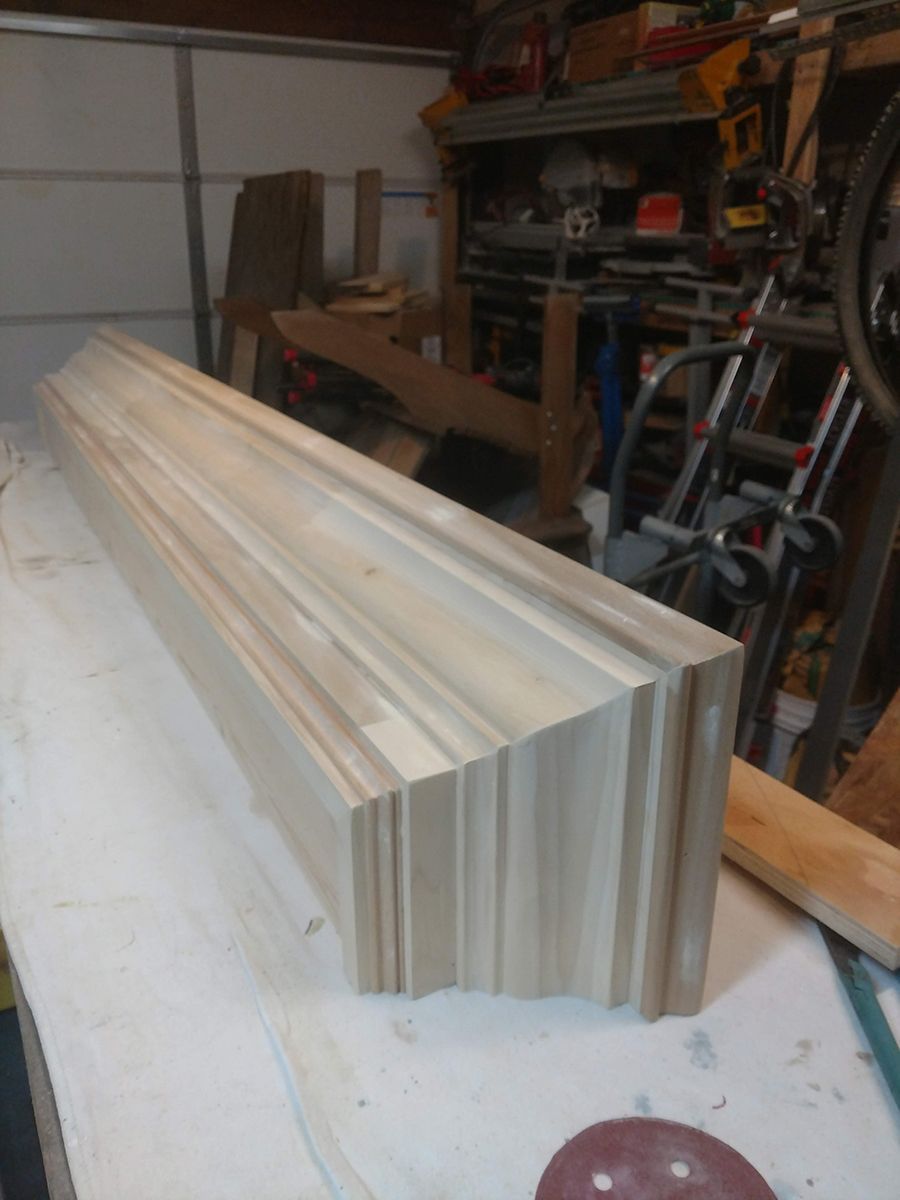 A wooden mantelpiece in a workshop. It is light-colored, angular, and resting on a work surface.