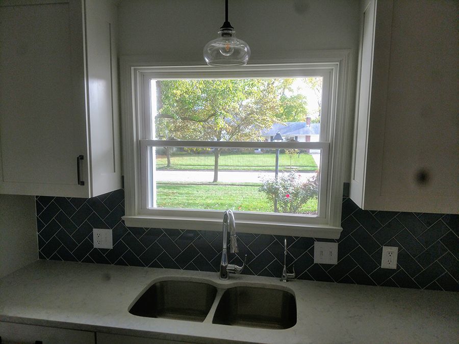 Kitchen with double sink, window, pendant light, dark blue backsplash, and white cabinets.