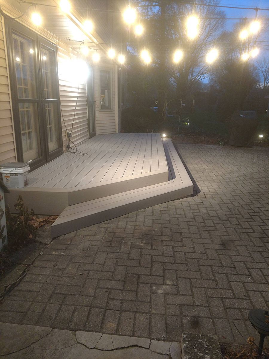 A wooden deck with steps, connected to a brick patio, lit by string lights.