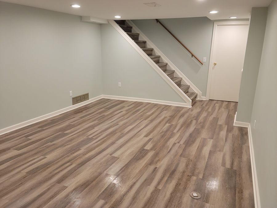 Empty basement room with light wood-look flooring, stairs, white trim, and a closed door.