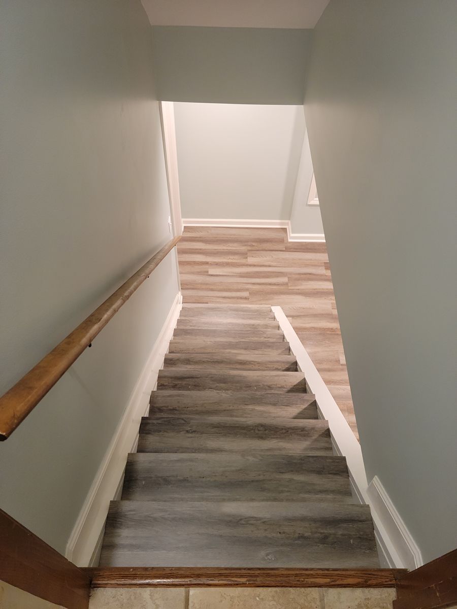 Staircase leading downwards; gray steps, wooden handrail, pale blue walls, wood-look flooring at the bottom.