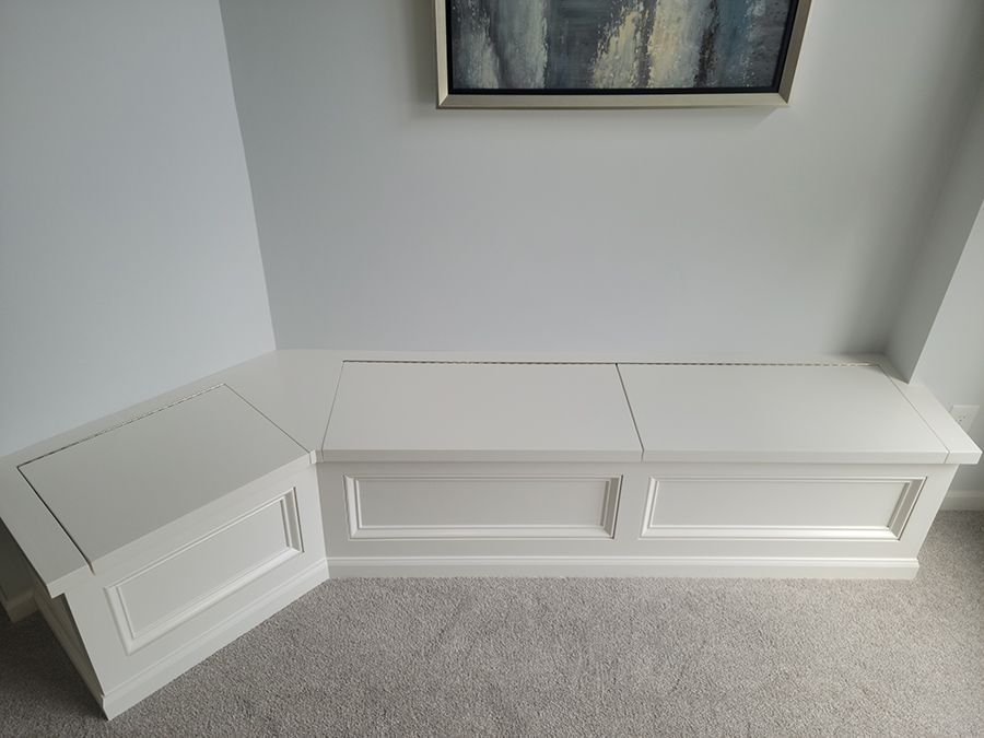 White built-in bench in a corner with a lifted lid, under a painting on a light gray wall, on a carpeted floor.