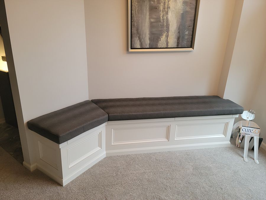 White built-in bench with gray cushion in a room with light walls. A framed abstract art piece hangs above it.