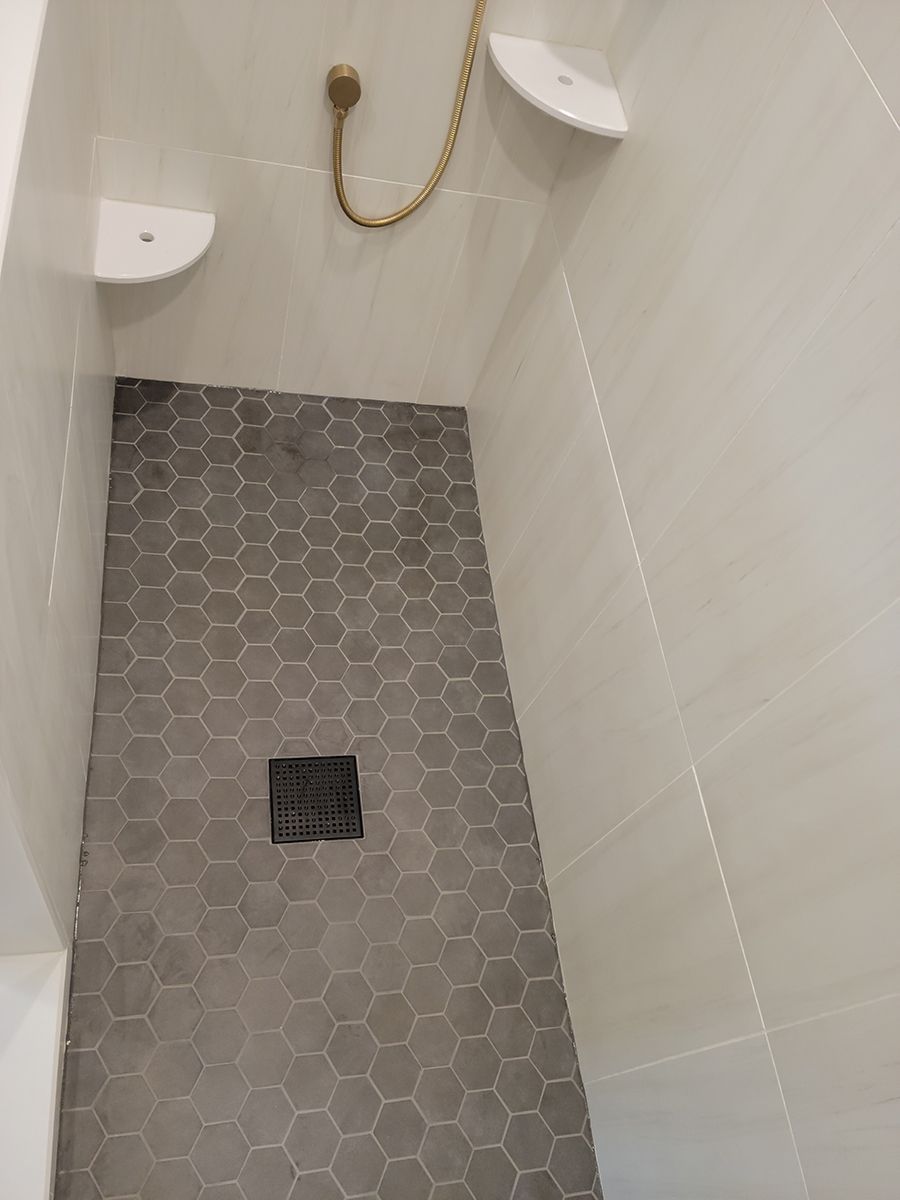 Shower with gray hexagon tile floor and white walls. A black drain is centered. Two white corner shelves are present.