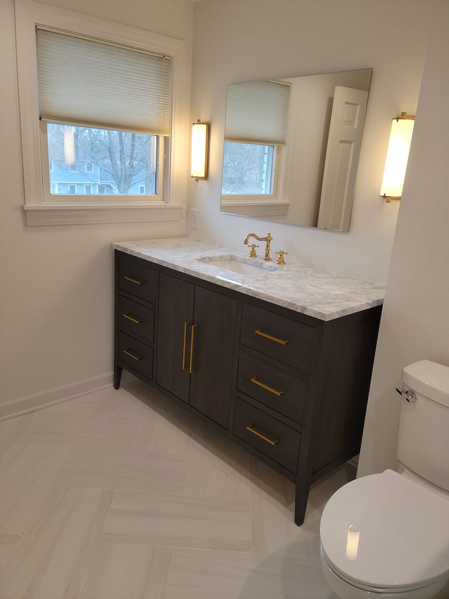 Bathroom with dark gray vanity, marble countertop, gold fixtures, and white tiled floor.