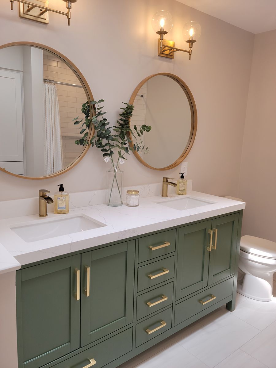 Bathroom with green vanity, gold fixtures, round mirrors, and greenery.