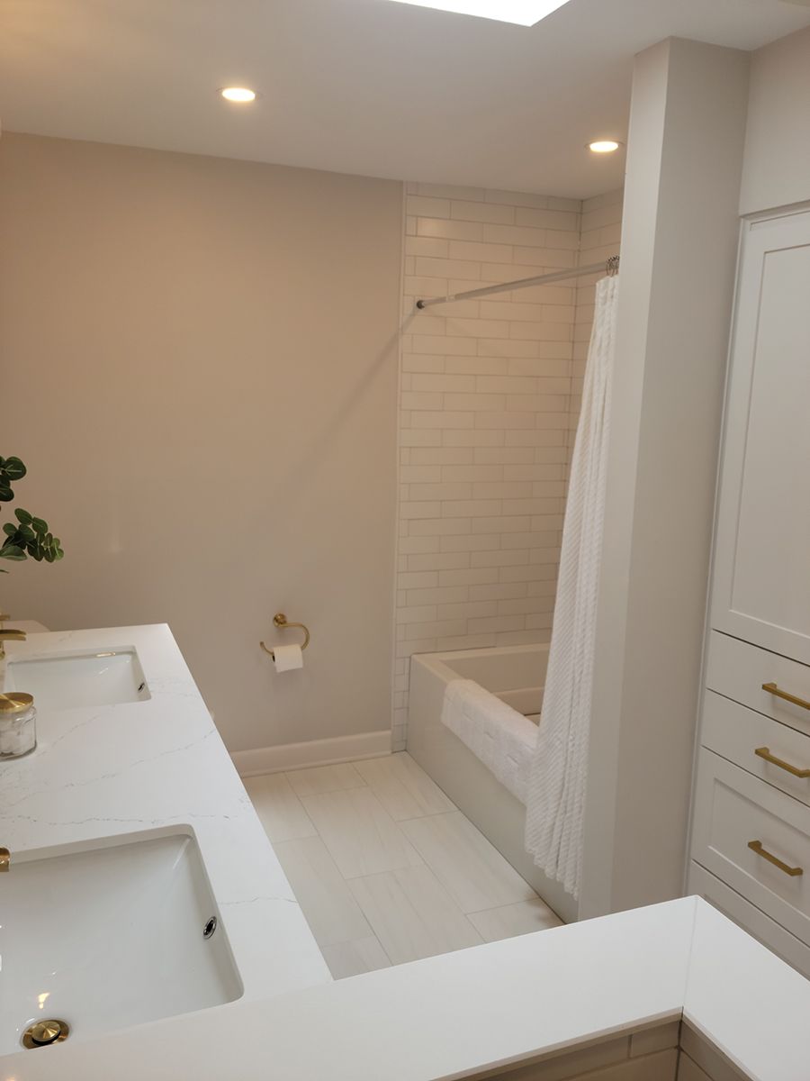 Bathroom with white sink, tub, and cabinets. Beige walls, skylight, and gold hardware.