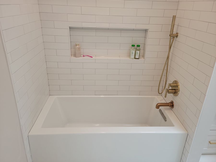 White bathtub with gold fixtures in a tiled alcove; toiletries in a recessed shelf.