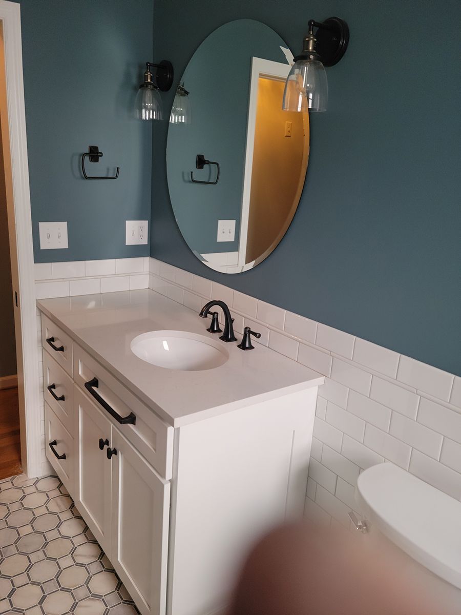 Bathroom with white vanity, oval mirror, blue walls, black fixtures, and white tiled backsplash.