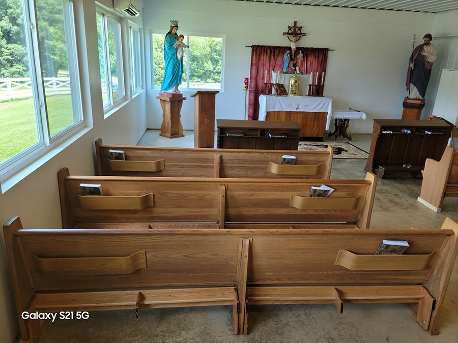 Interior of a small chapel with wooden pews, statues, an altar, and large windows overlooking greenery.