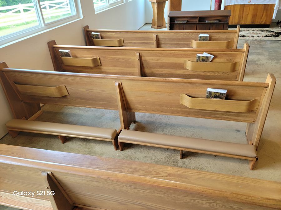 Wooden church pews with tan cushions. Some pews have booklet holders. Indoors, near a window.