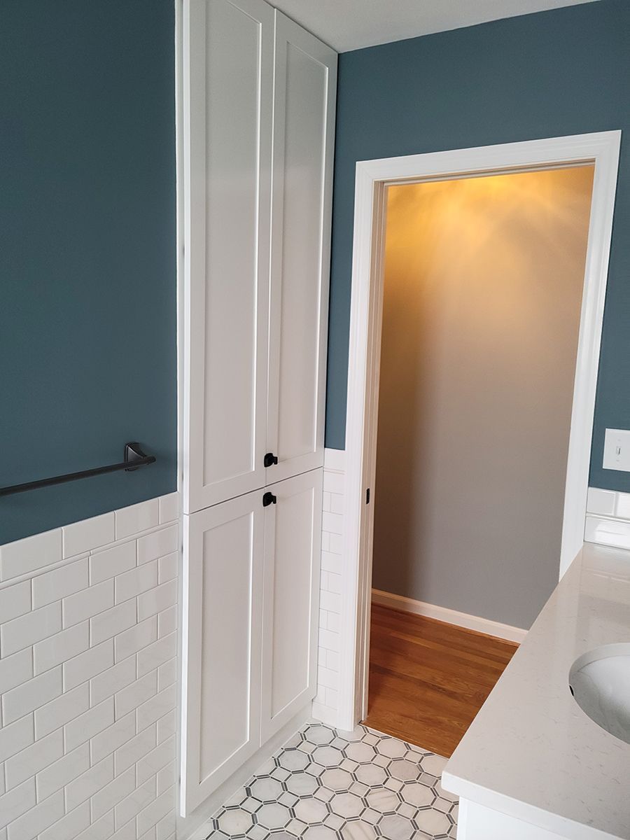 White tall cabinet in a bathroom, blue wall, white tile, doorway to another room.