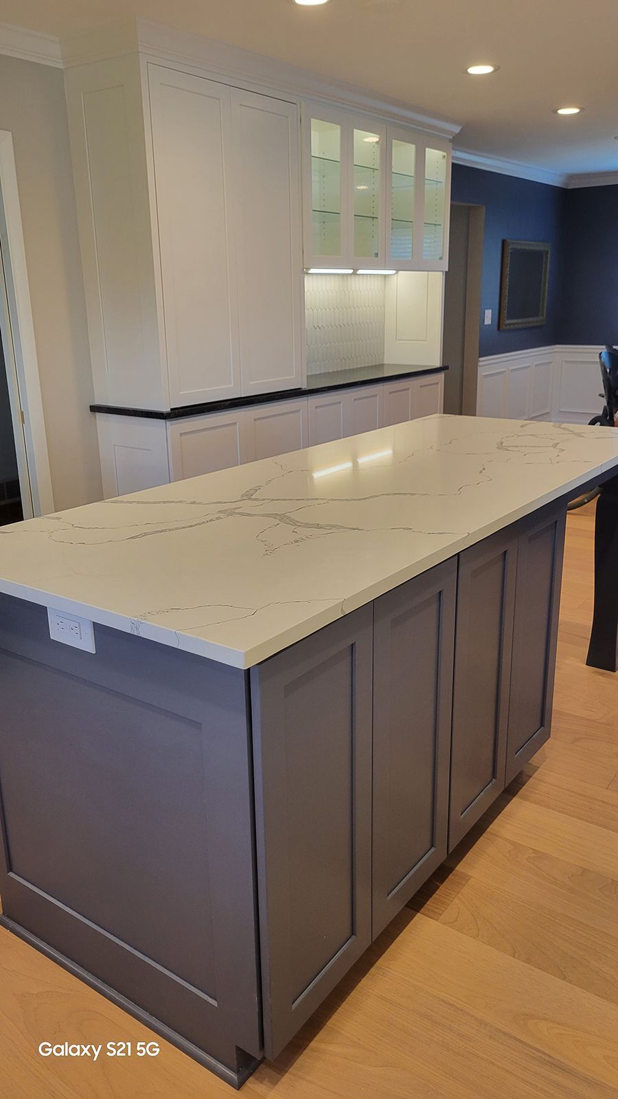 Gray kitchen island with white countertop. Light-colored wooden floor. Cabinets in the background.