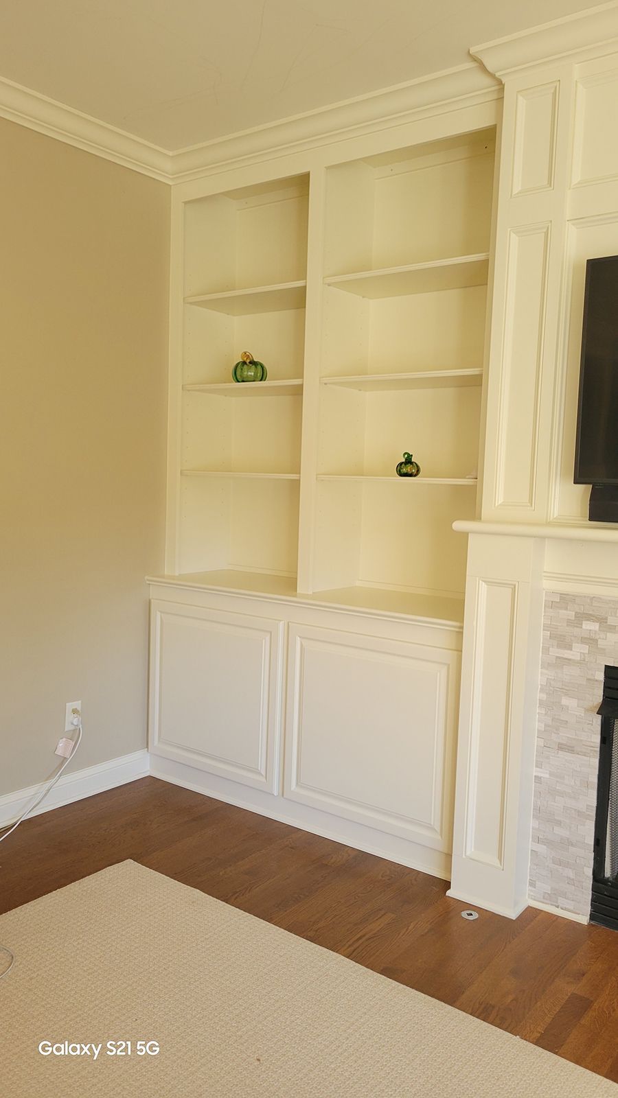 Built-in white bookshelves with upper open shelves and lower cabinets, adjacent to a fireplace with a light-colored rug and wood floor.