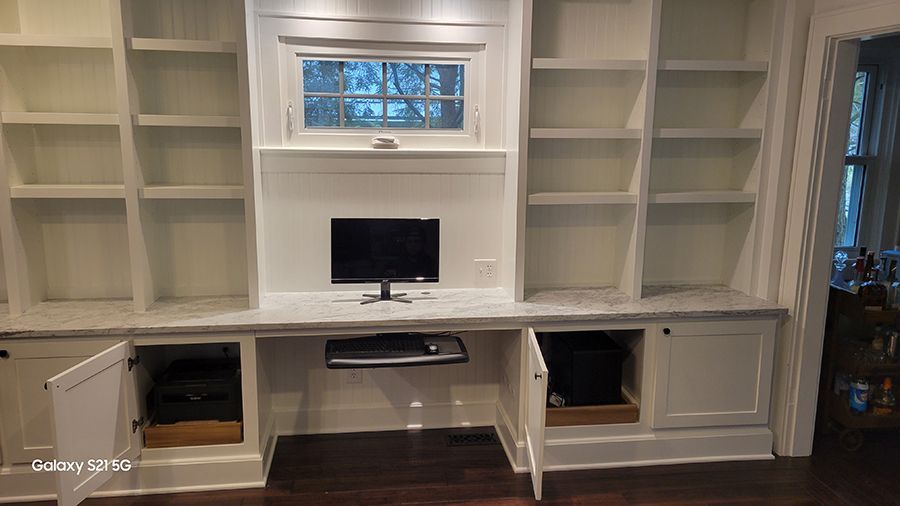 White built-in shelving unit with desk area. A computer sits on the desk. Open lower cabinets reveal storage.