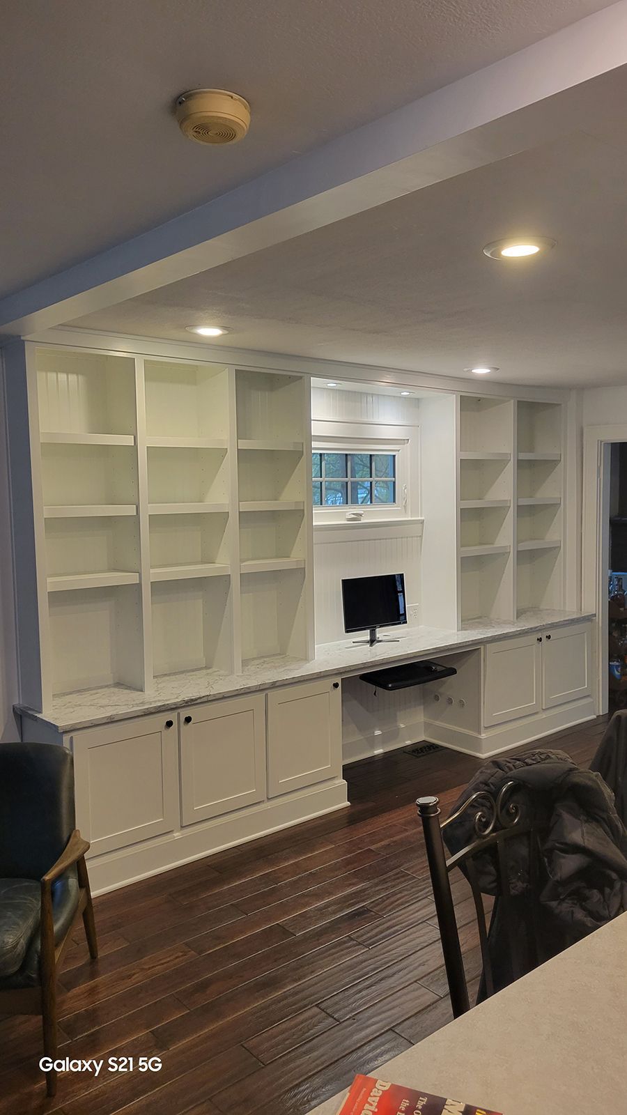 Built-in white bookshelves and cabinets with a desk area, installed against a wall with dark wood flooring.