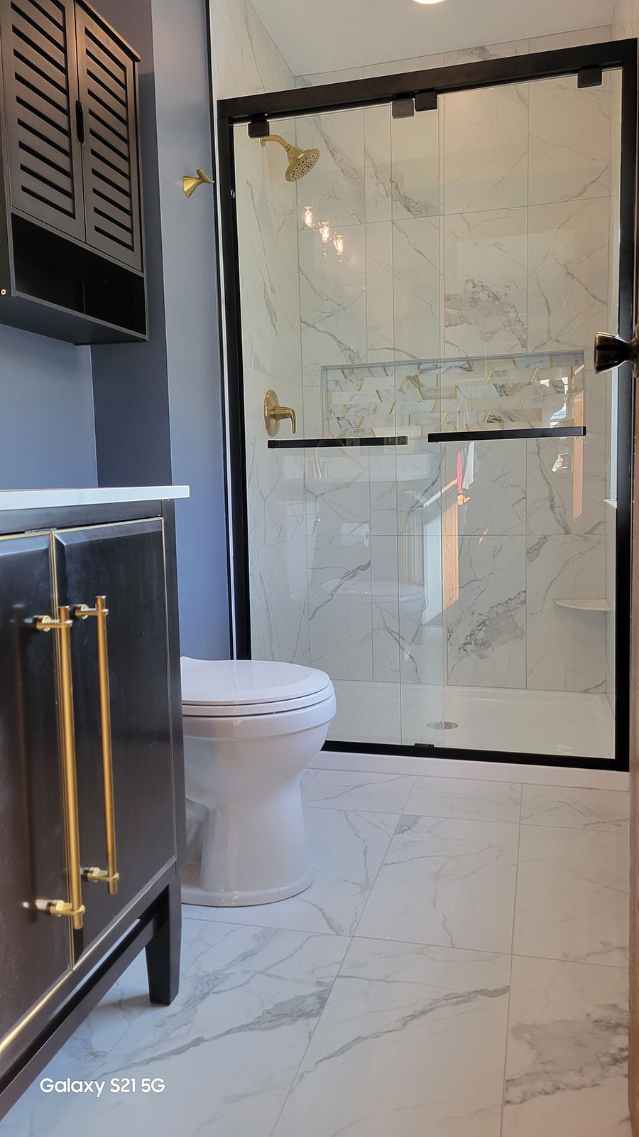 Modern bathroom with white marble tile, glass shower, and dark cabinetry with gold hardware.