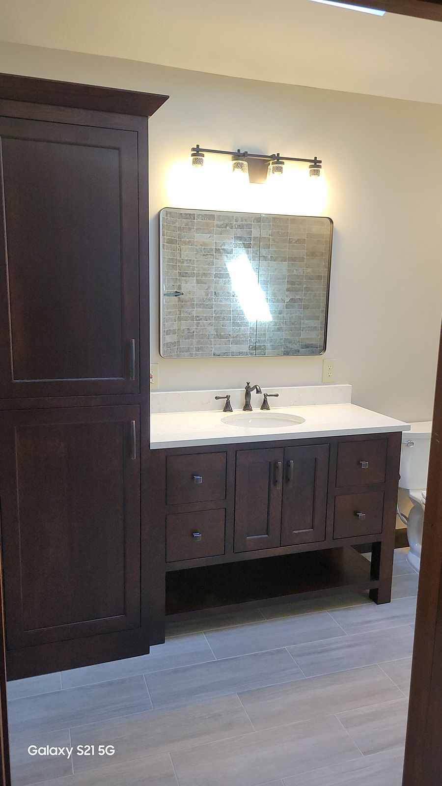 Bathroom with dark brown vanity, tall cabinet, stone-look mirror, and light fixtures.