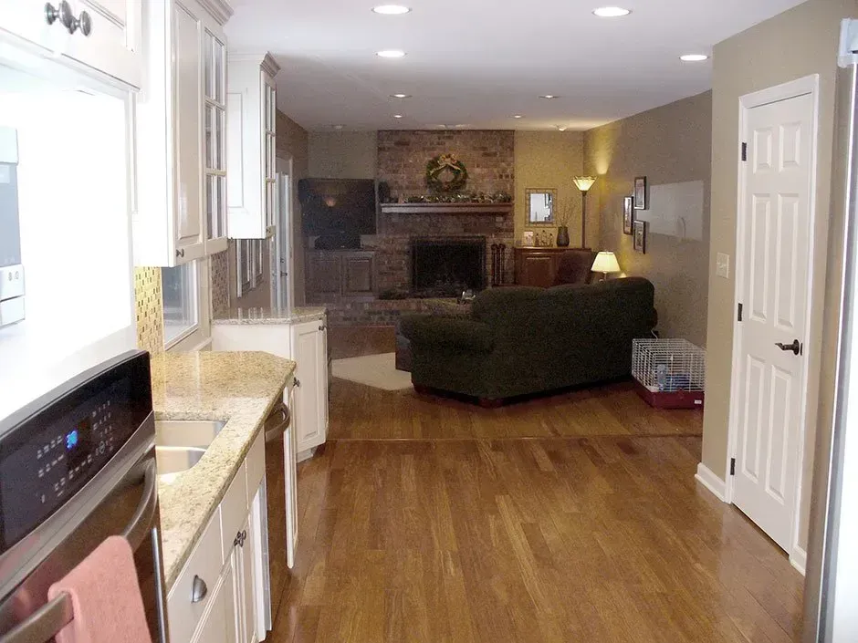 Kitchen with light cabinets and granite countertops, opening to a living room with a fireplace and dark green sofa.