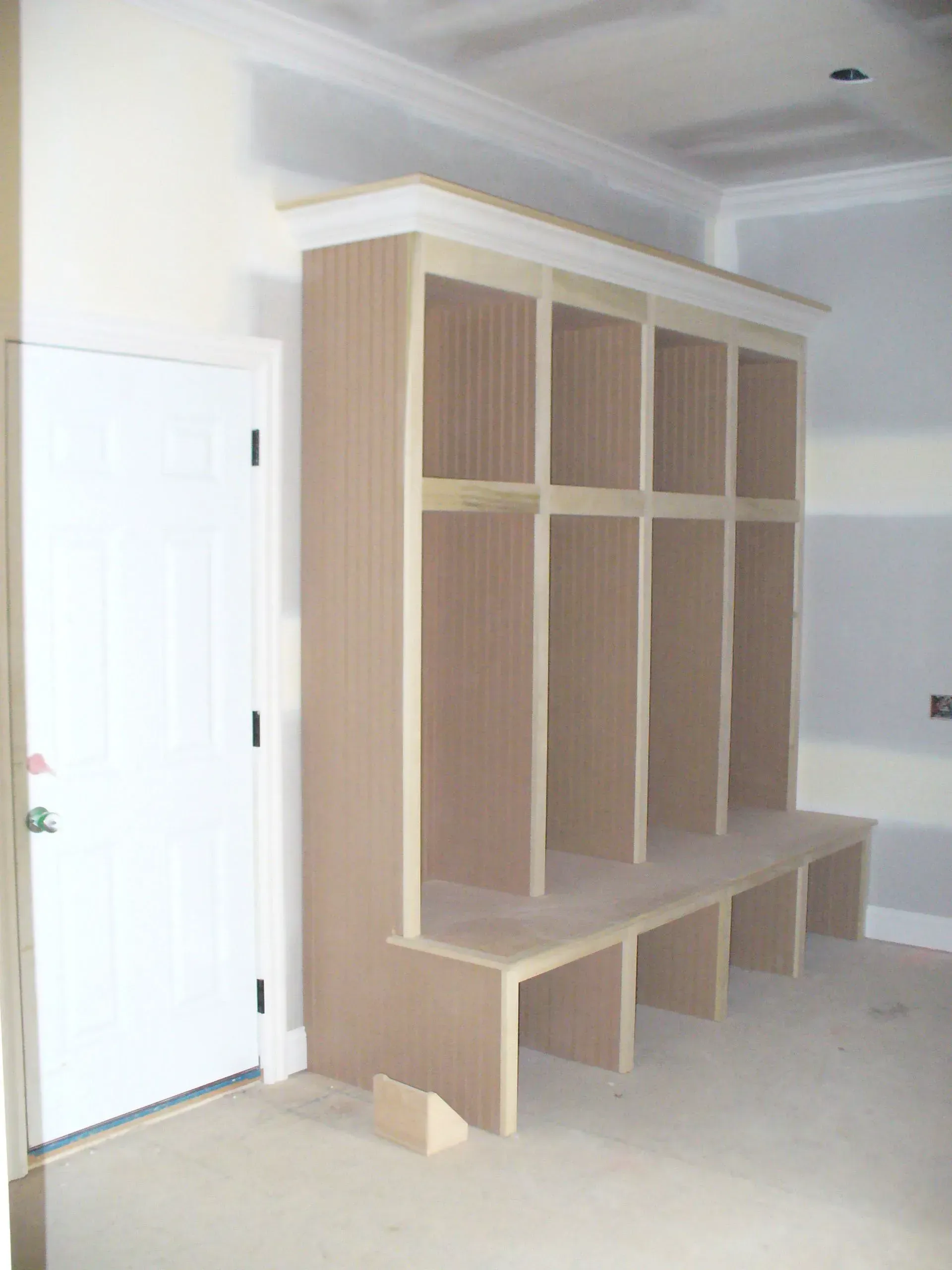 Unfinished beige wooden built-in bench with cubbies and trim, next to a white door in a room under construction.