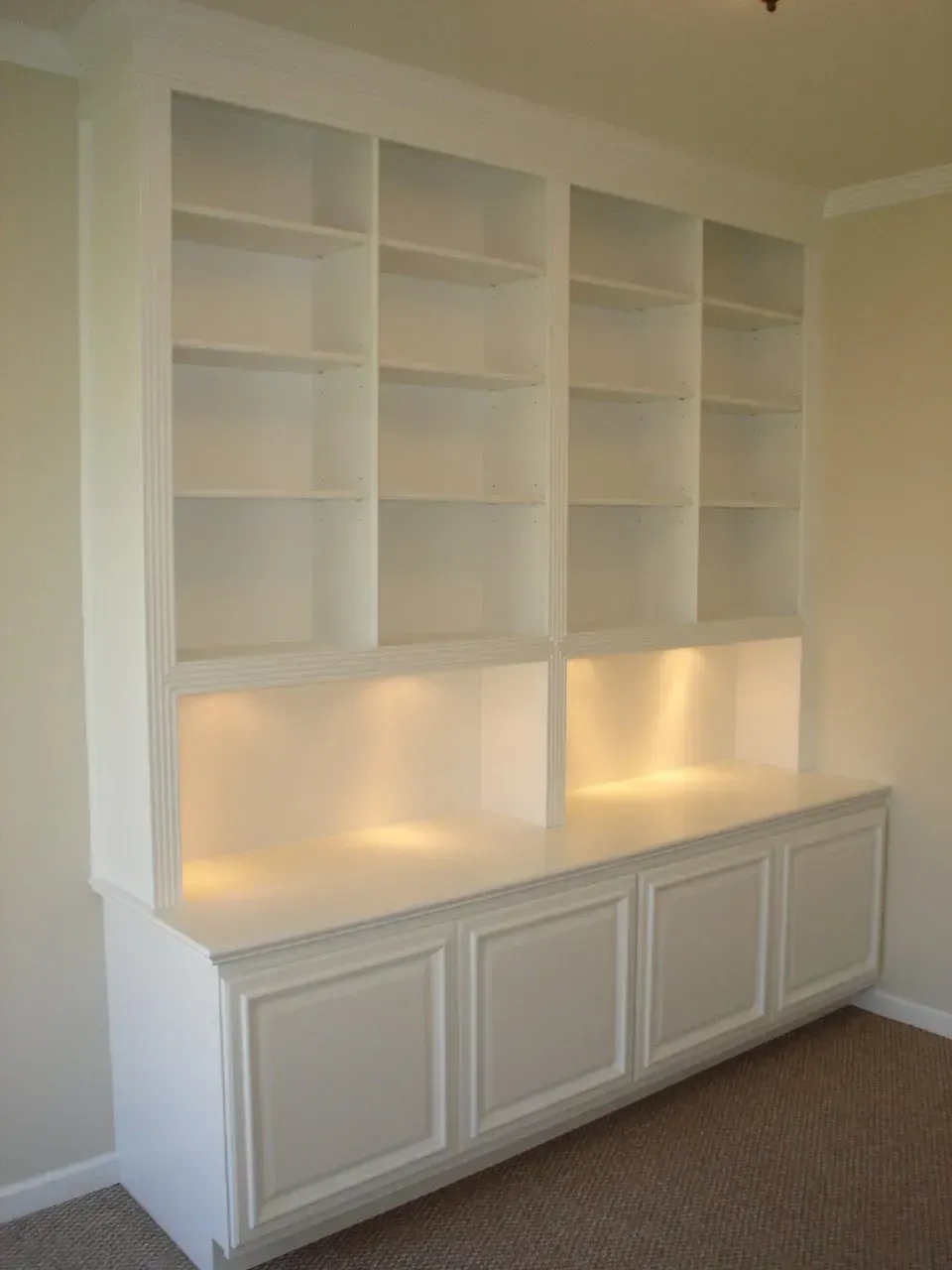 Built-in white bookcase with shelves, lower cabinets, and interior lighting. Set against a light beige wall.