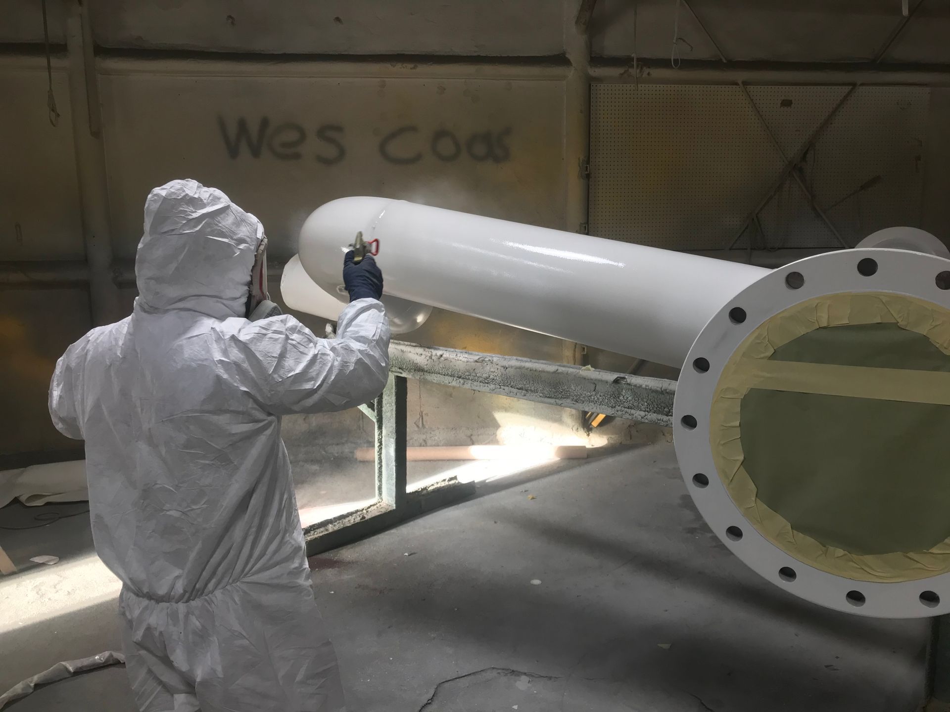 Worker in white protective suit spray-painting a large white pipe in an industrial workshop