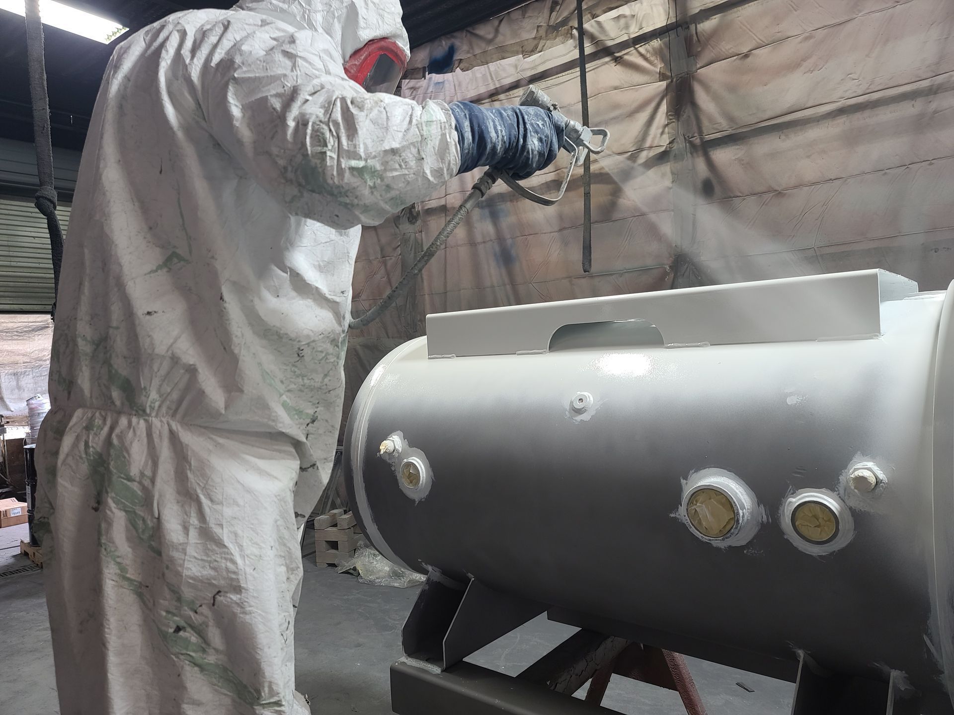 Worker in white protective suit spray-painting a gray cylindrical metal tank in an industrial workshop