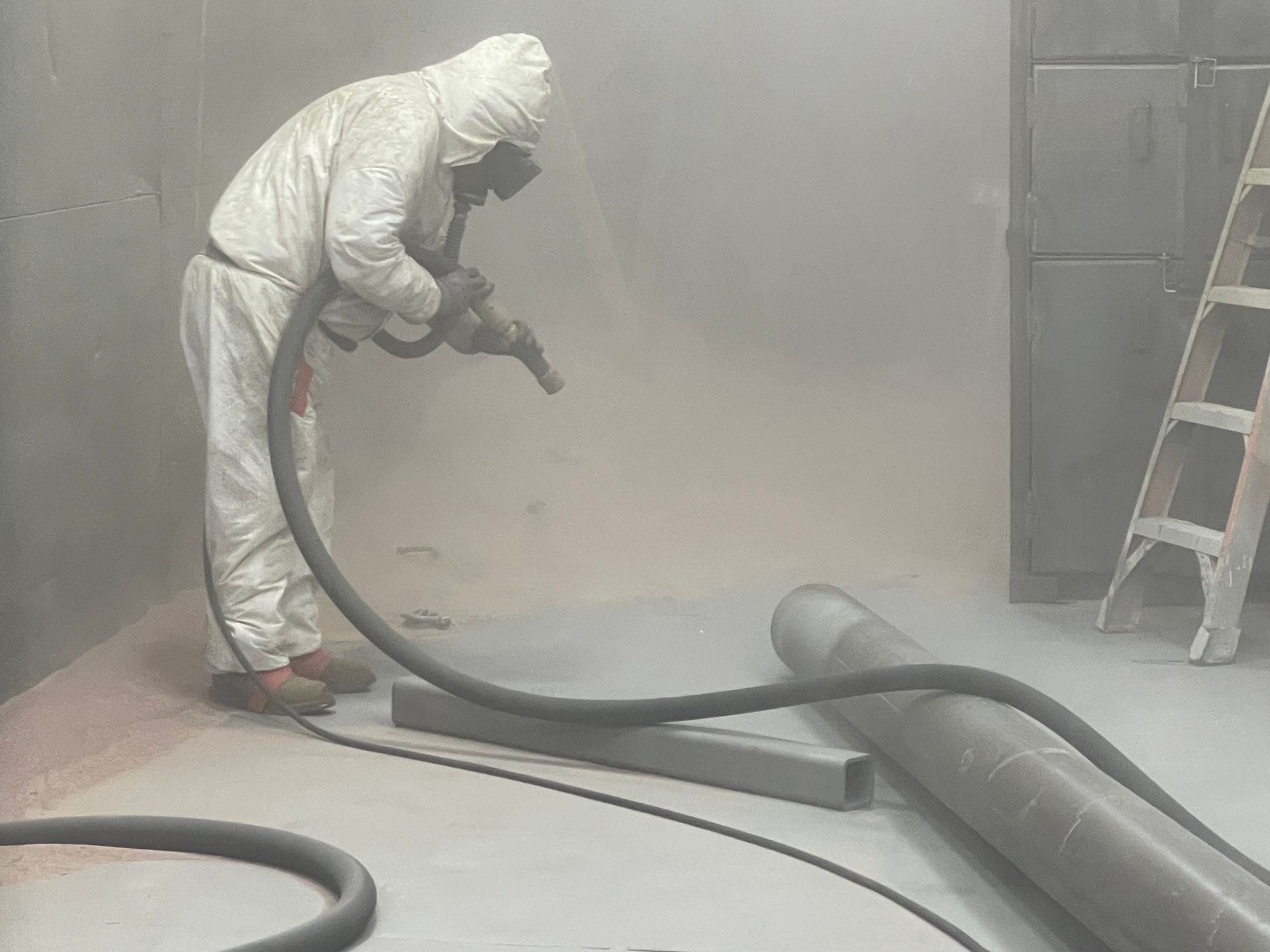 Worker in protective suit vacuuming dust in a hazy room beside a ladder and large pipe