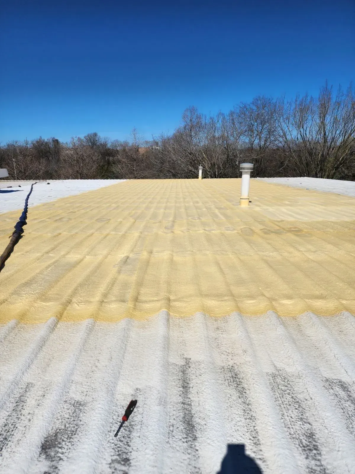 A flat roof with yellow coating, vents, and tool; blue sky and trees in the background.