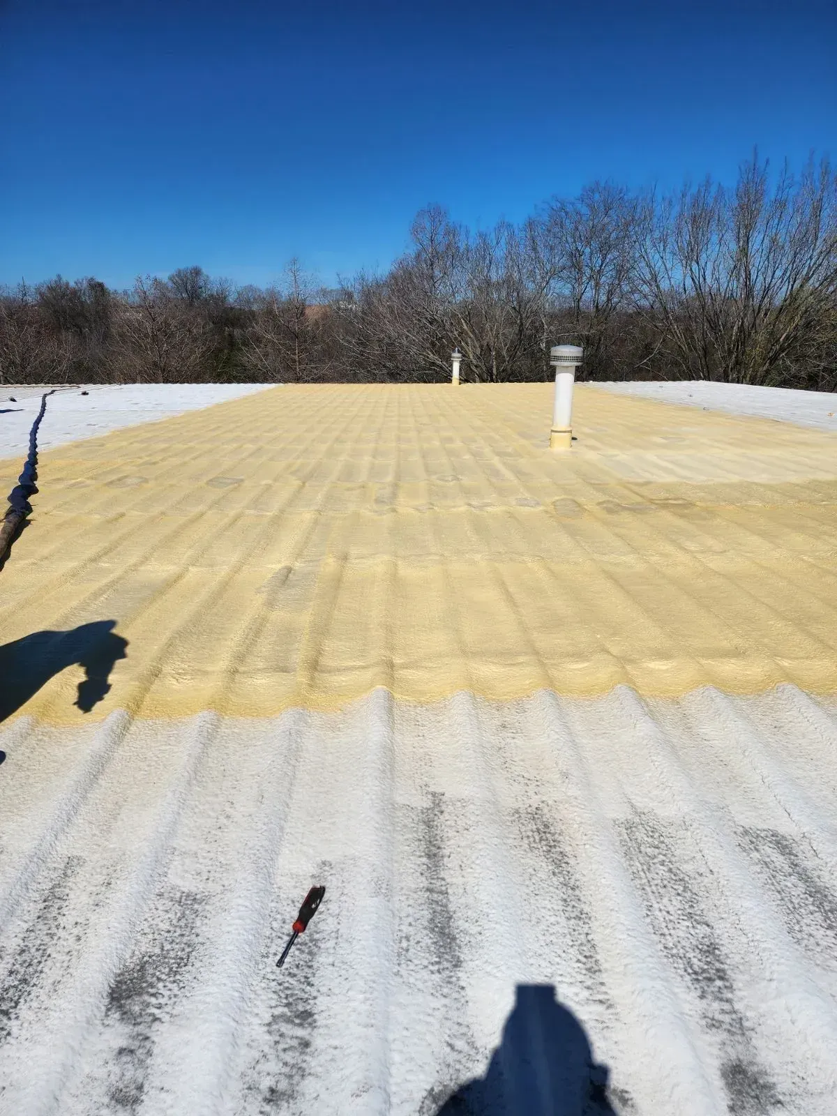 Yellow foam insulation on a corrugated metal roof, with a clear blue sky and trees in the background.