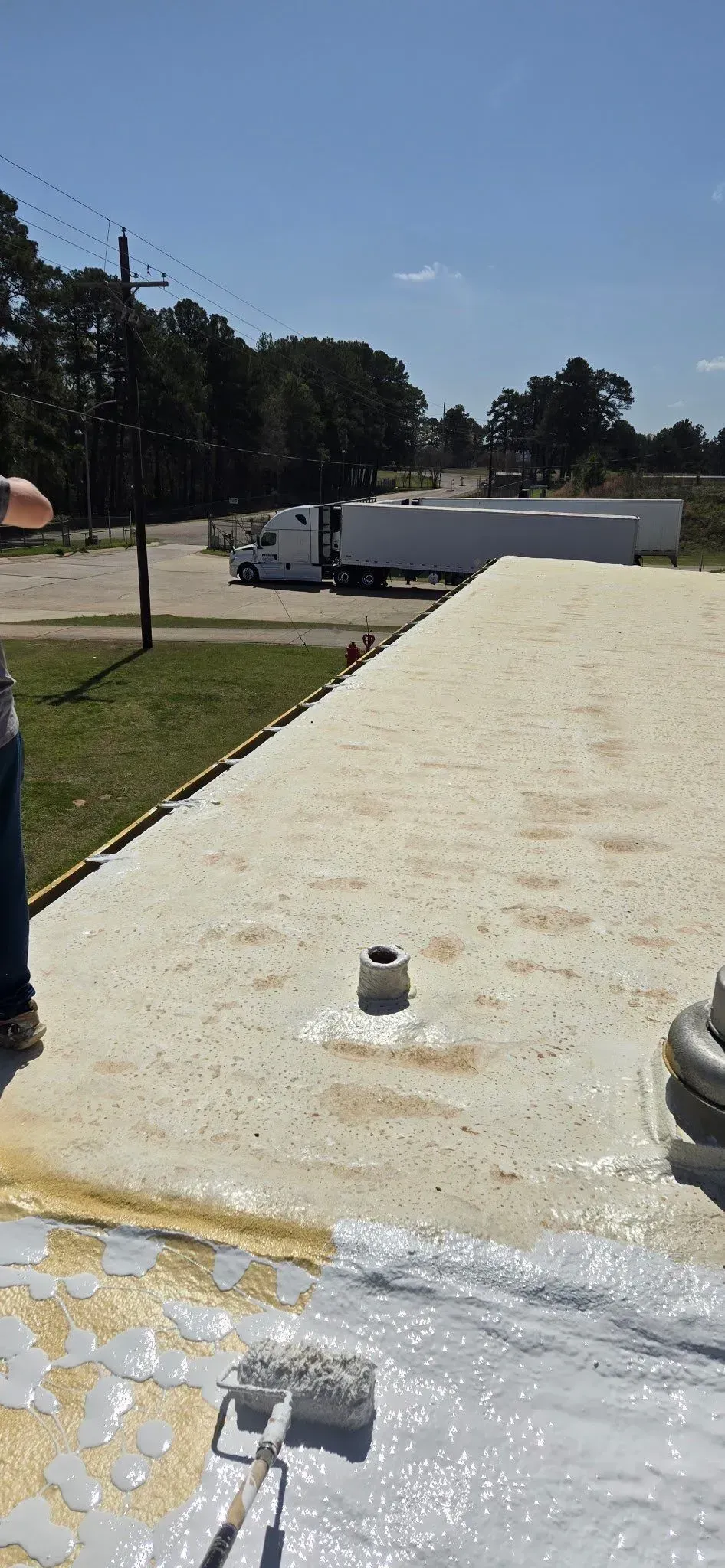 A person applies a sealant to a roof; a semi-truck is visible in the background on a sunny day.