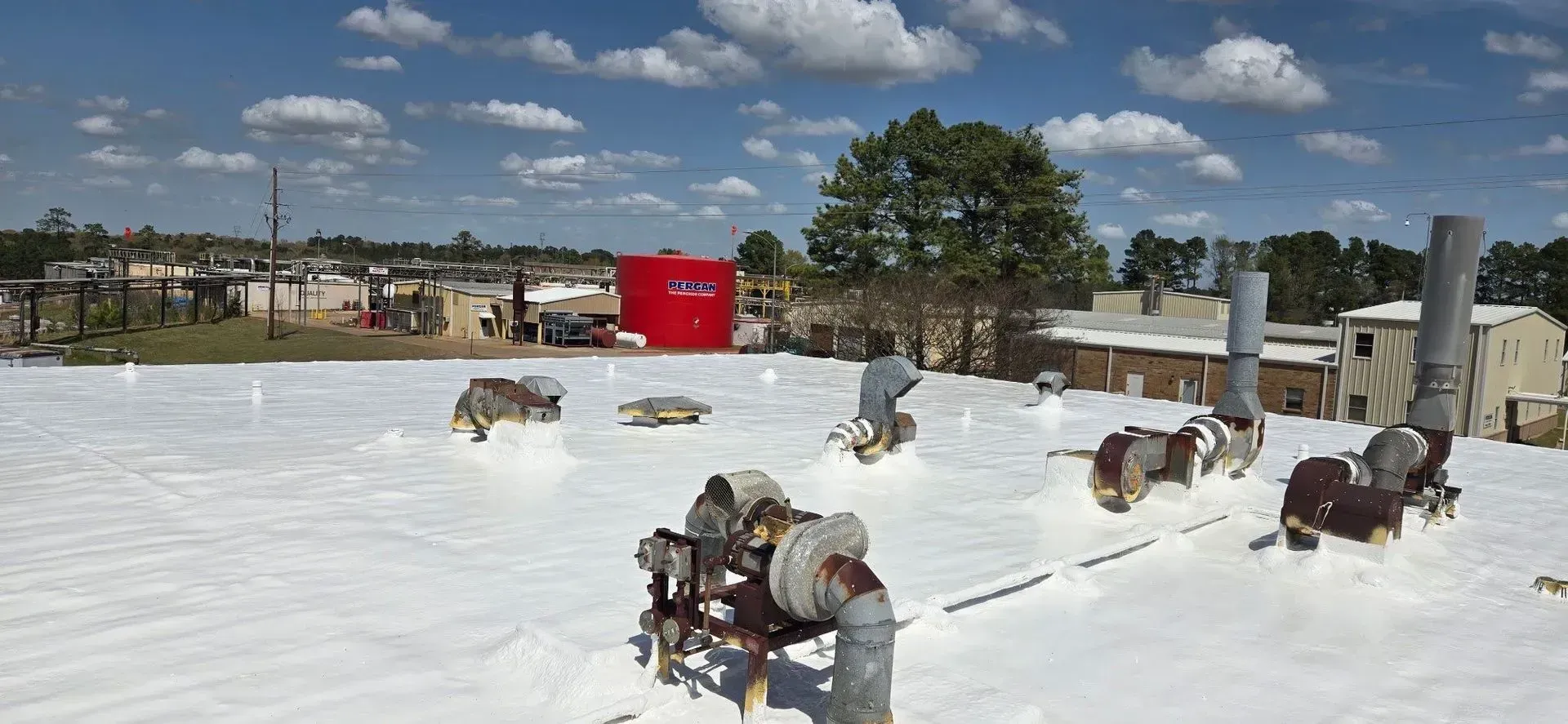 A roof covered in white material with ventilation pipes and machinery. Buildings and trees in background.