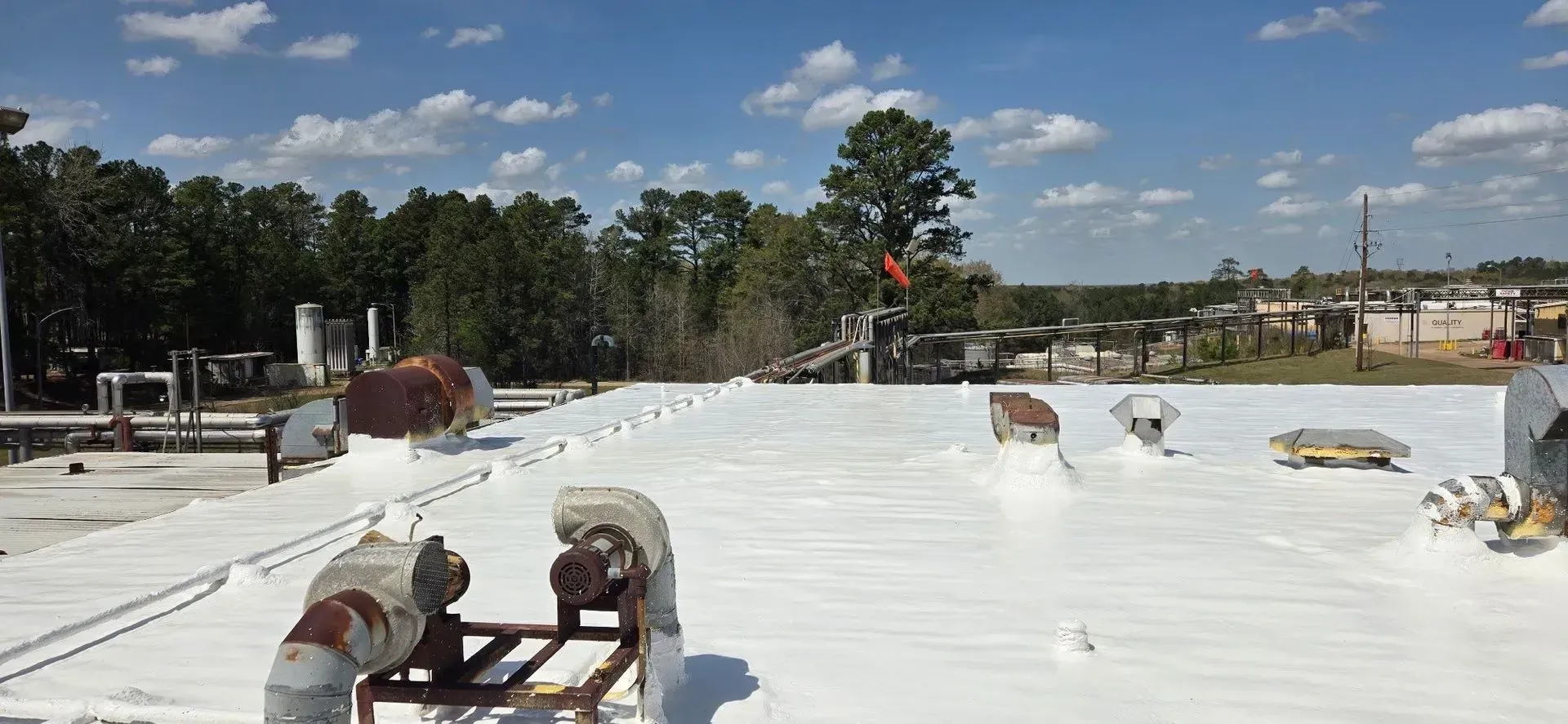 A white-coated roof with equipment, trees in the background, and a bright blue sky.