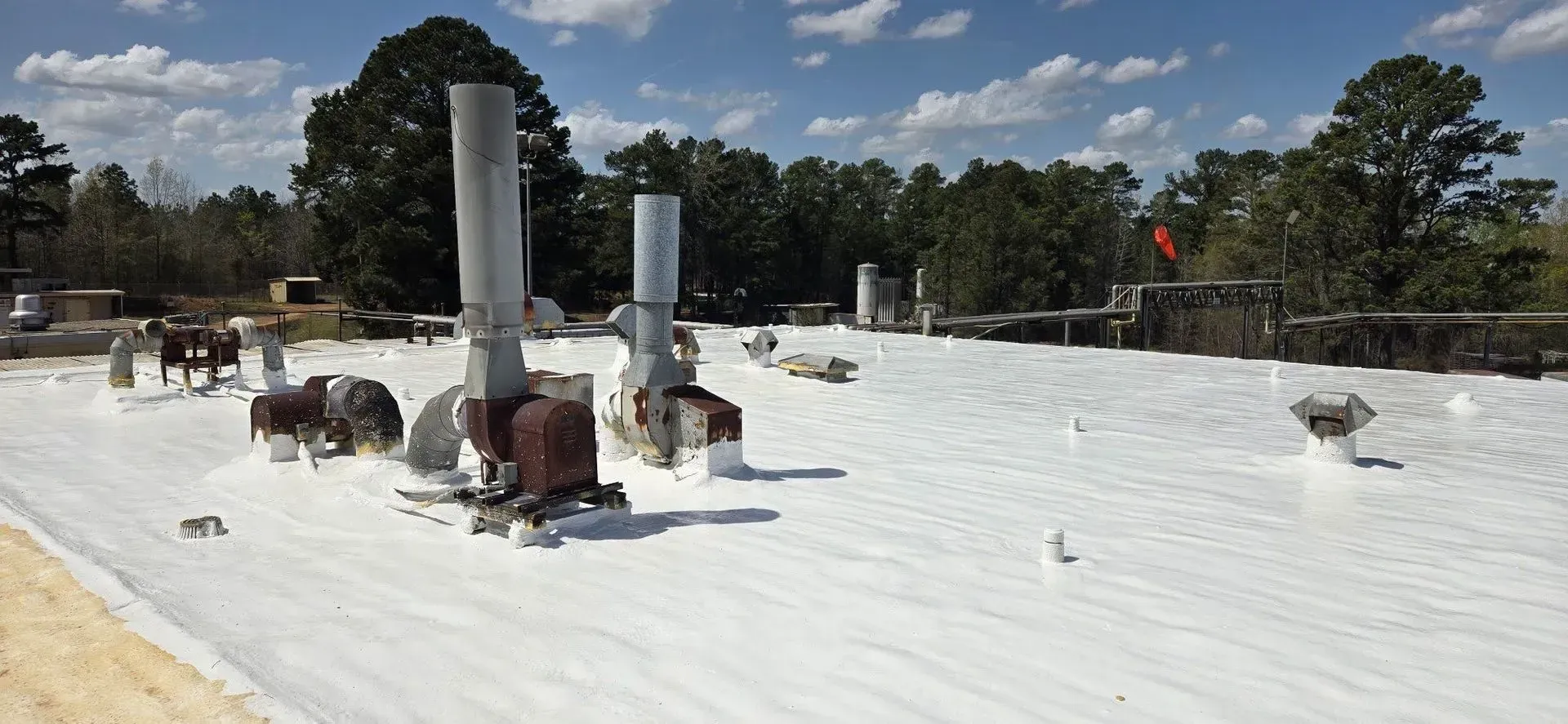 A flat white roof with several vertical pipes and figures; trees and blue sky with clouds in the background.