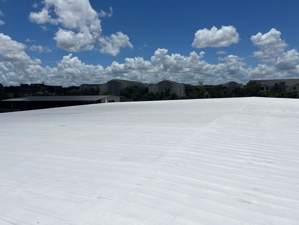 A wide, bright white flat commercial roof under a sunny blue sky with scattered clouds and buildings in the background.