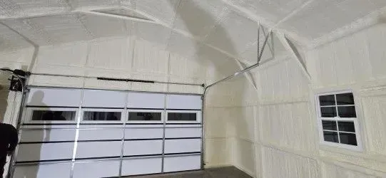 Interior of a garage insulated with white spray foam. A closed garage door is visible, and a small window is on the right.