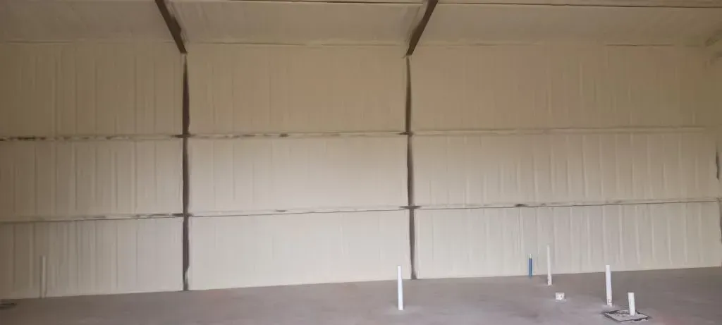 An empty, insulated warehouse interior. Beige walls, gray floor, white pipes protrude.