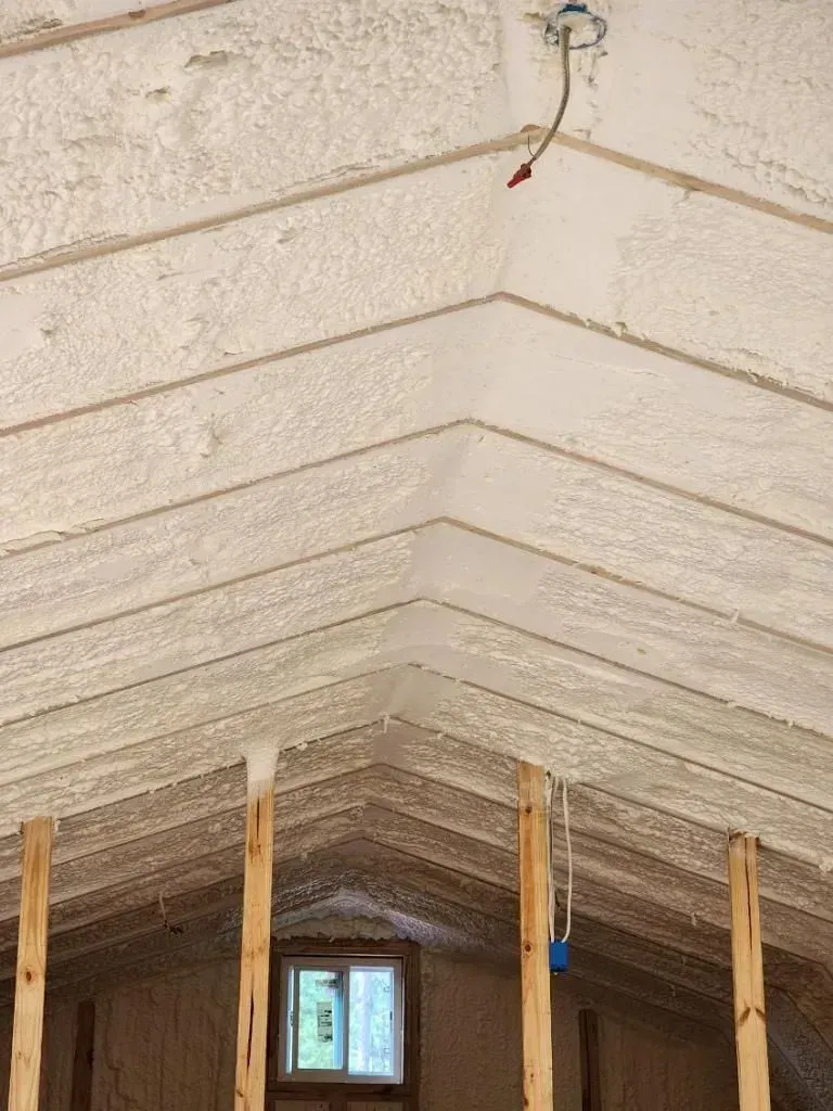 Angled ceiling with exposed wood beams and spray foam insulation. Electrical fixture visible.