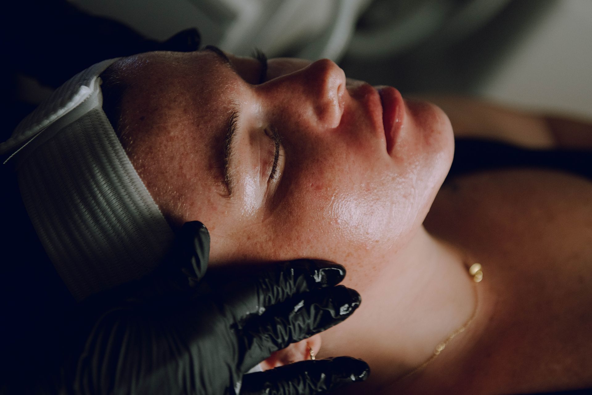Person receiving facial treatment, hands in black gloves gently touch face. White headband, spa setting.
