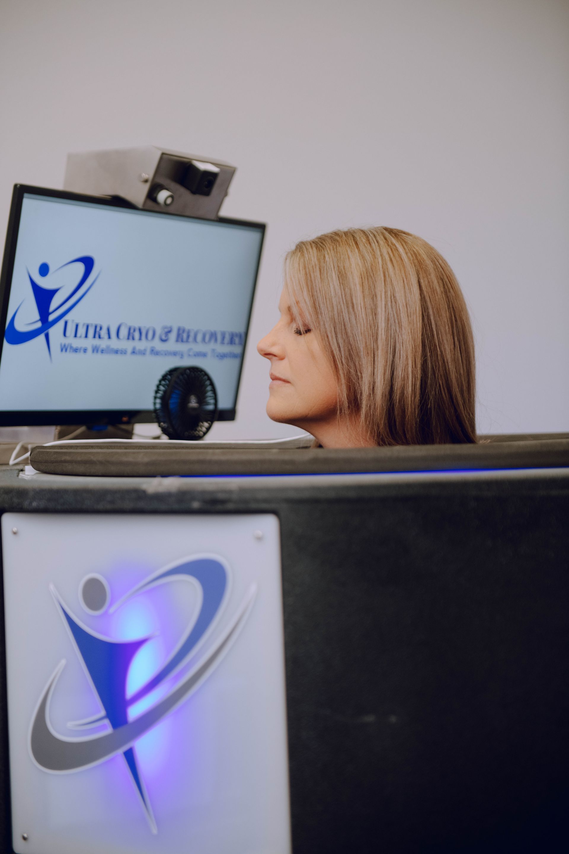 Woman with head in recovery device, facing screen with logo. Gray, blue, white.
