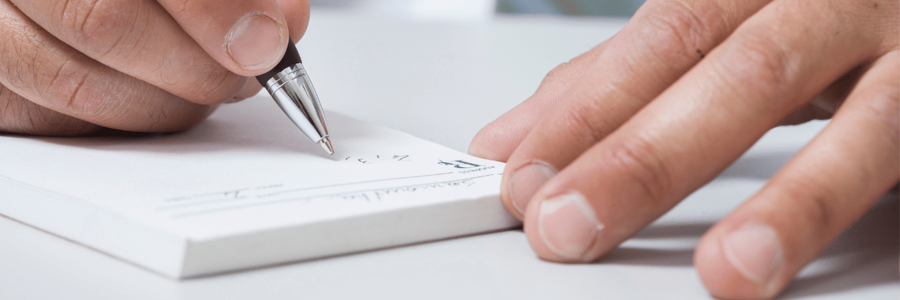 A person's hand writing on a white notepad with a pen. Another hand rests on the notepad to stabilize it.
