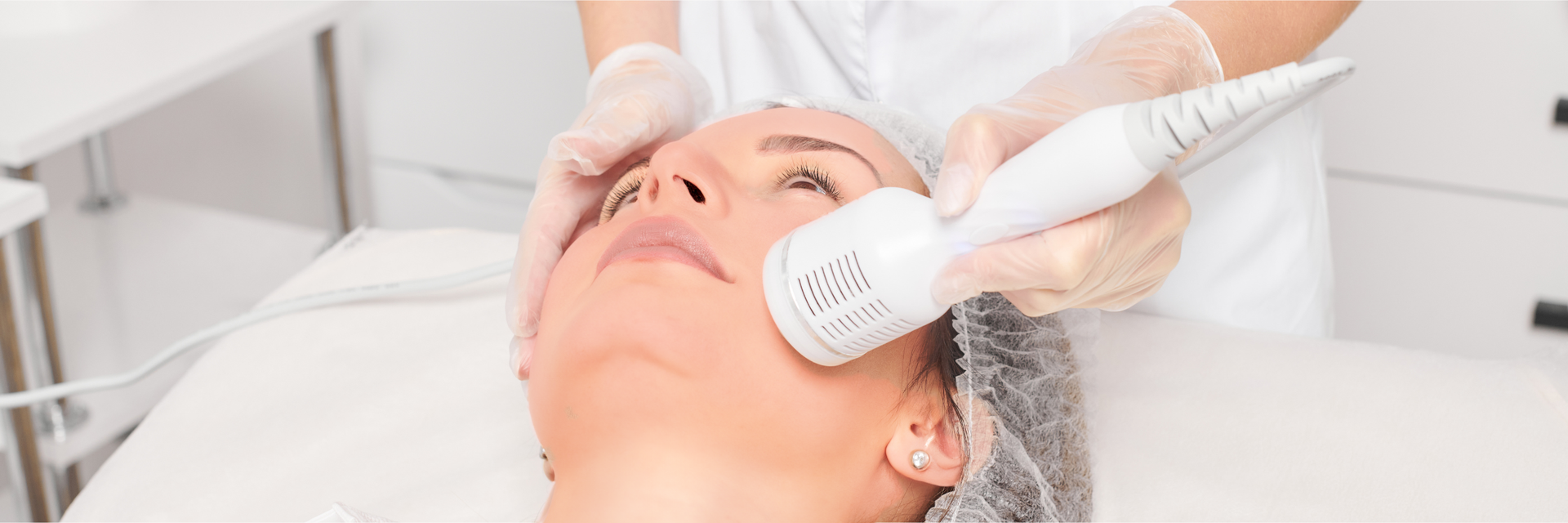 Facial treatment with a device; hands in gloves. Woman lying down, looking up. White setting.