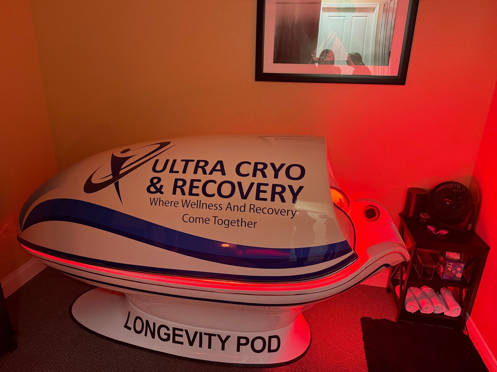 An Ultra Cryo & Recovery longevity pod with white and blue branding sits in a dimly lit room bathed in red light.