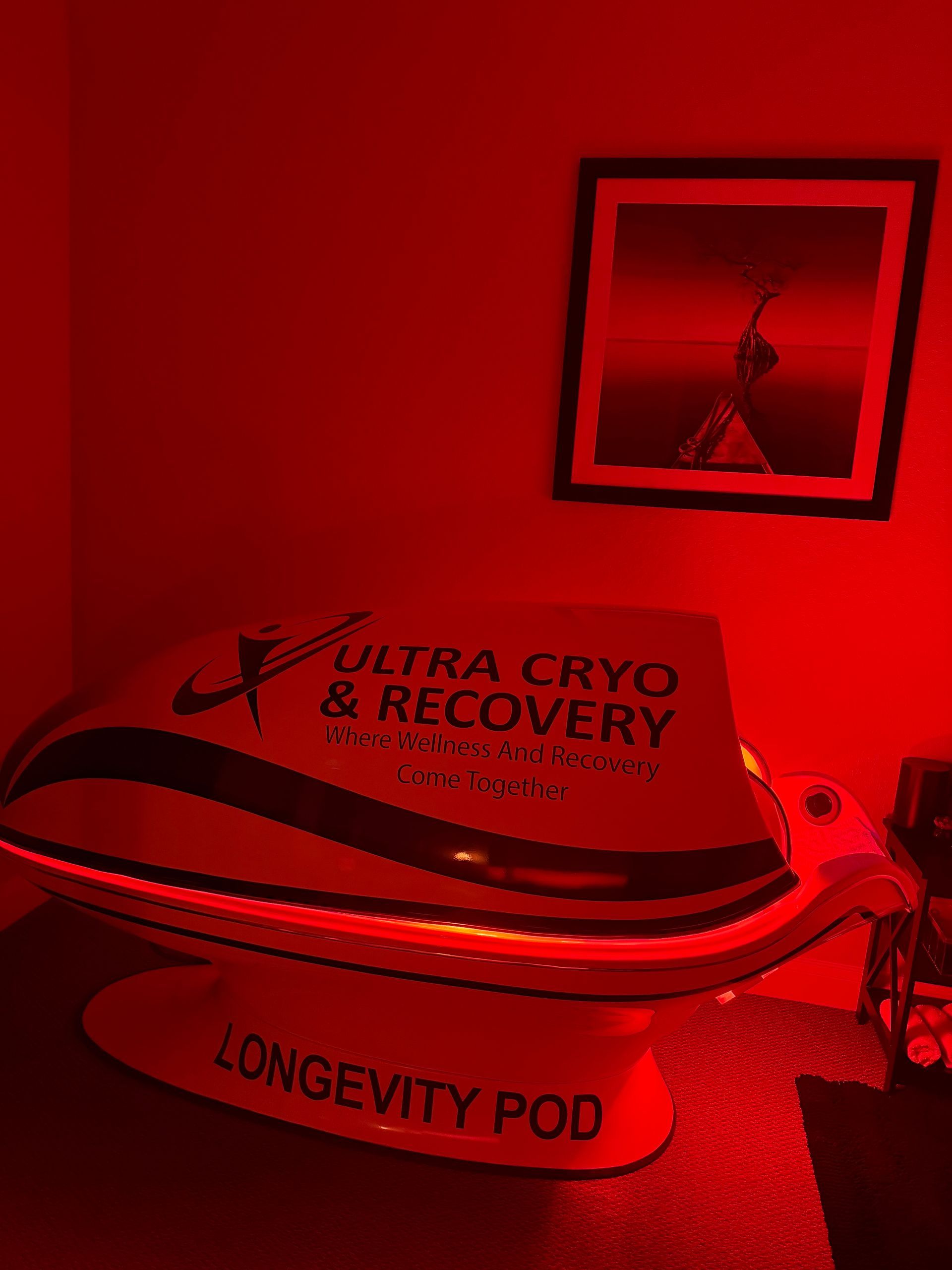 A Longevity Pod by Ultra Cryo & Recovery sits in a dim room illuminated by red light, with framed art on the wall.