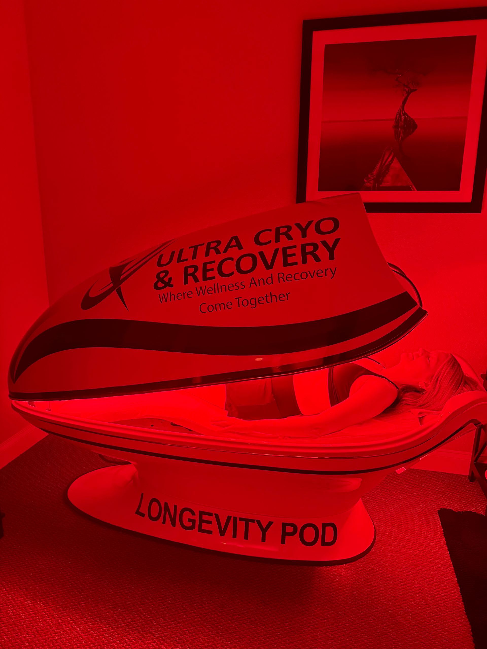 A white Longevity Pod recovery machine with red lighting in a room with a framed art piece on the wall.