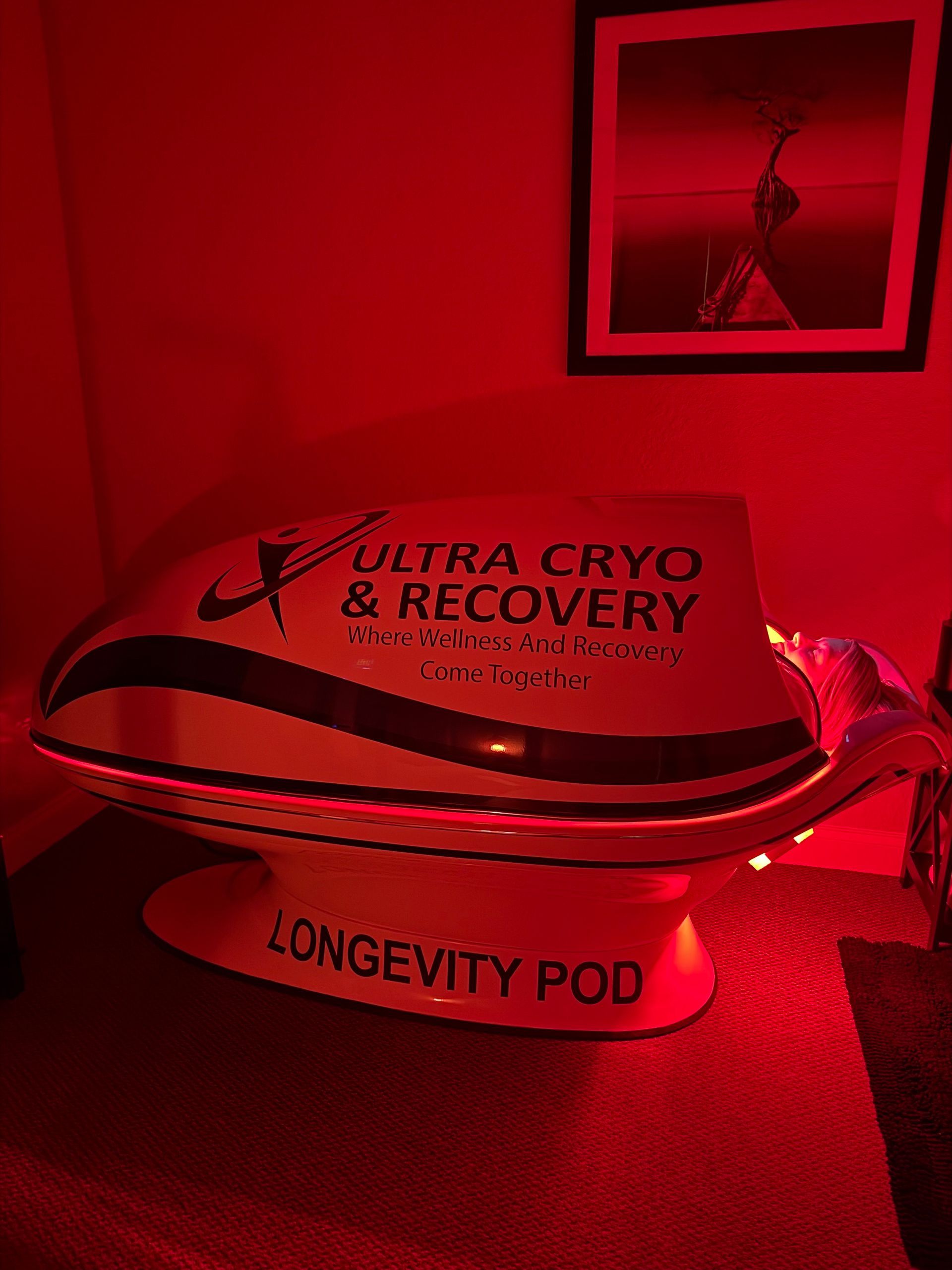 A white Longevity Pod by Ultra Cryo & Recovery sits in a dimly lit, red-toned room with wall art.