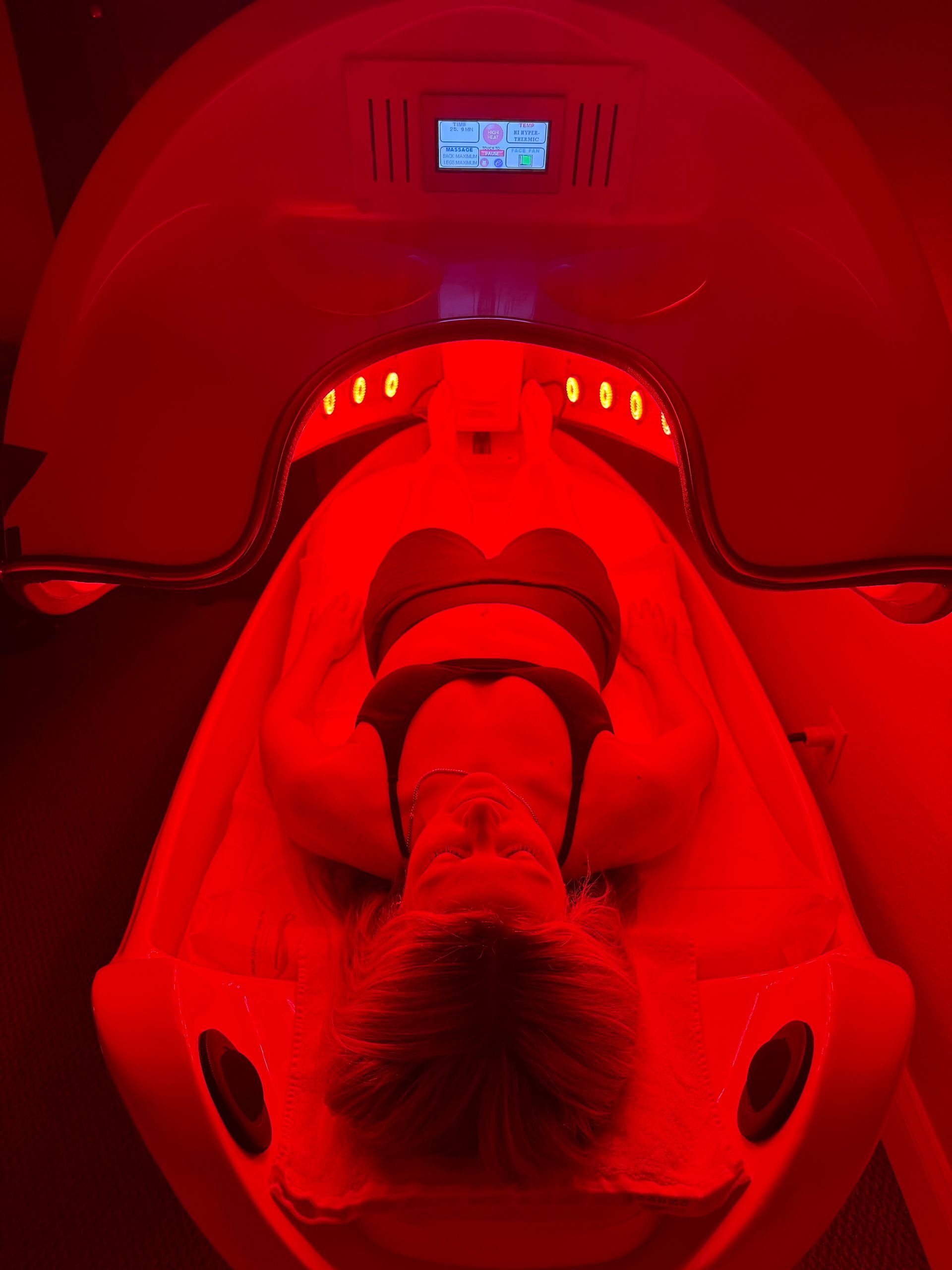 A person lies inside a red light therapy bed, with bright red light filling the chamber.