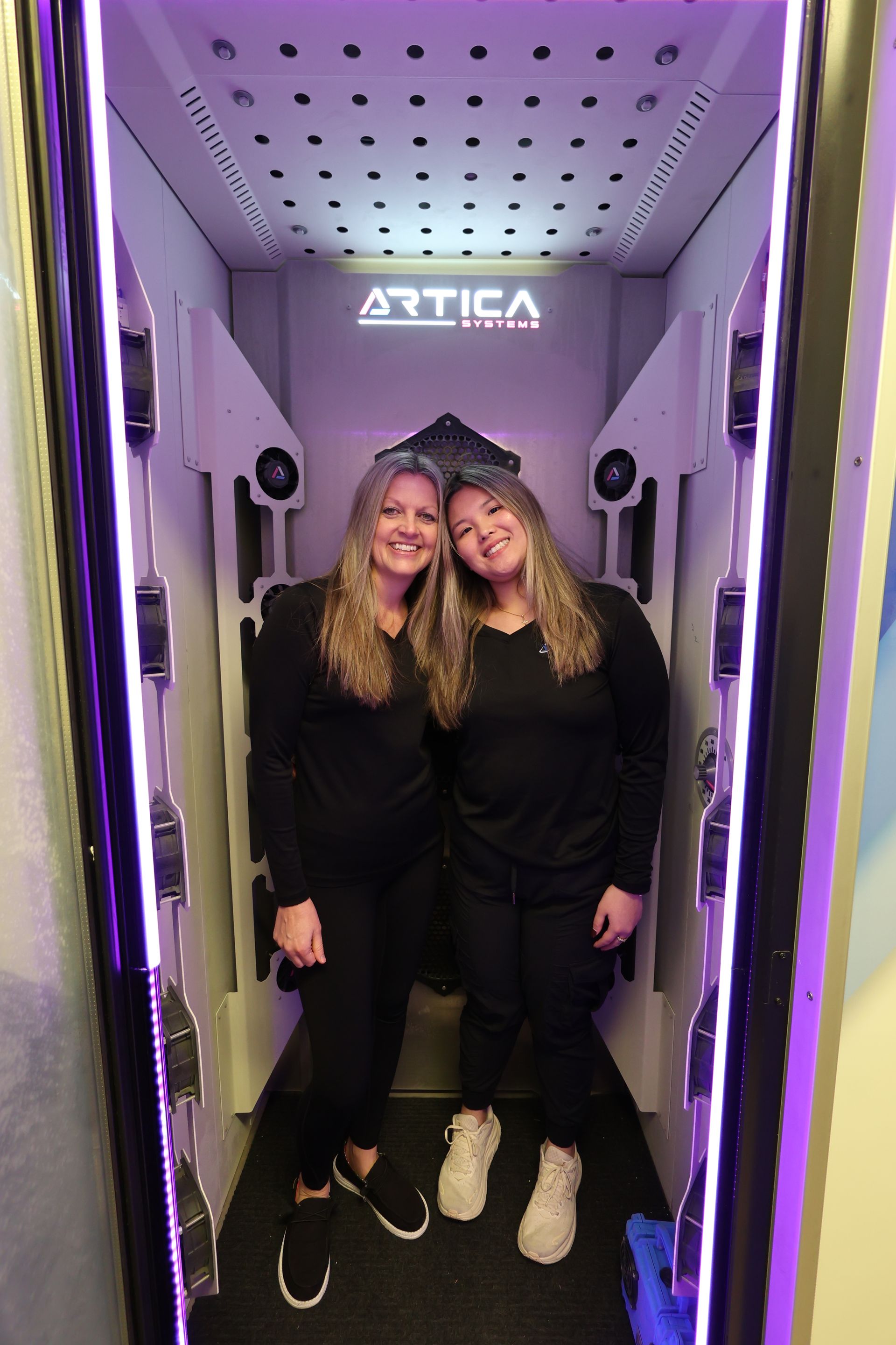 Two people stand smiling inside an Artica Systems cryotherapy chamber with white walls and bright purple side lighting.