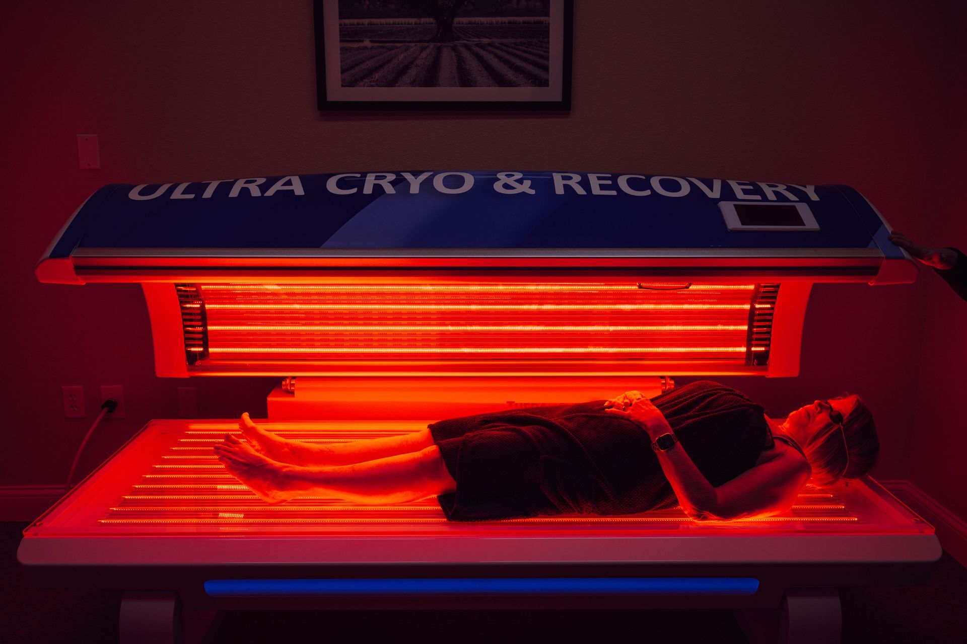 A person lies inside a red light therapy bed labeled Ultra Cryo & Recovery in a darkened room.