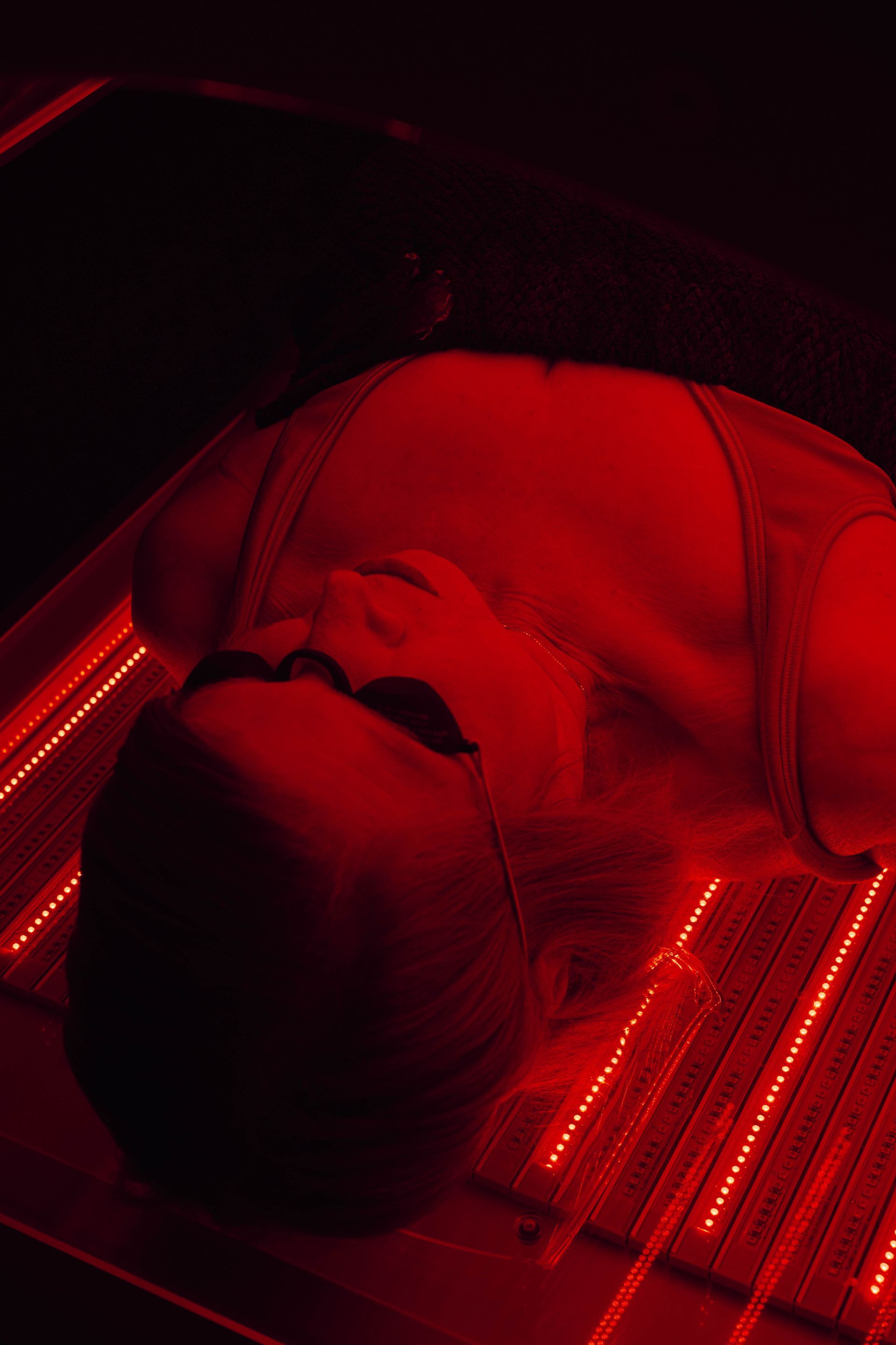 A person with sunglasses lies in a red light therapy bed under bright red LEDs.